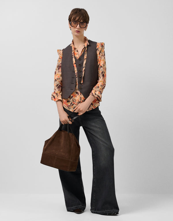 Person wearing a patterned shirt, vest, and wide-leg pants holding a brown bag on a white background