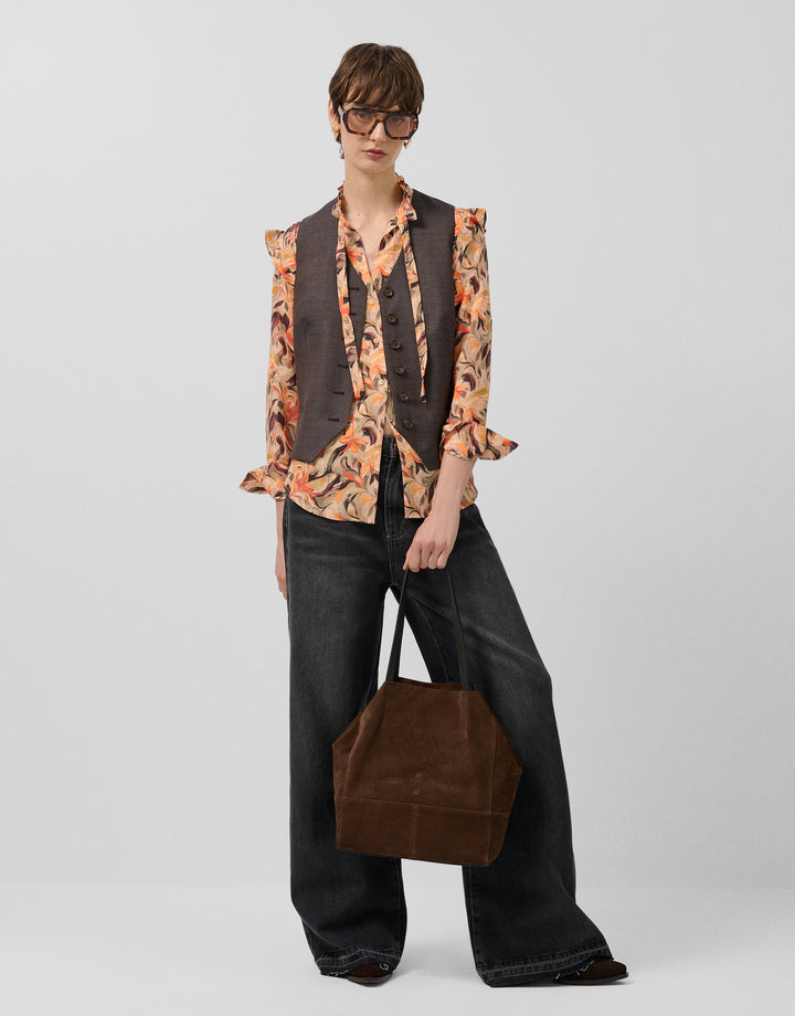 Person wearing a patterned blouse, vest, and black pants with a brown bag on a plain background