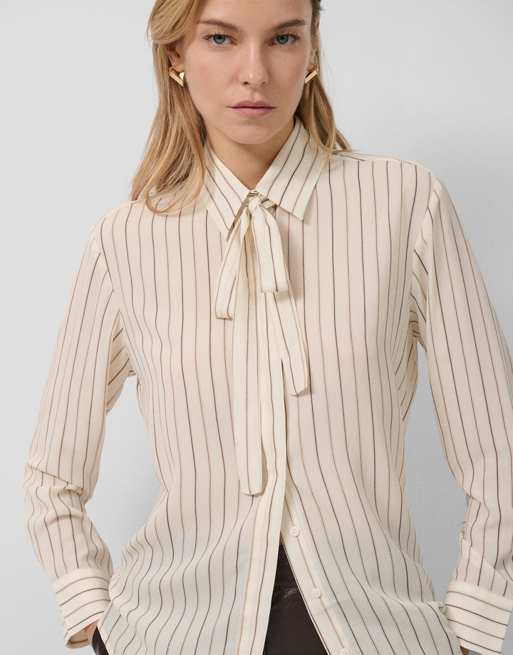 Woman wearing a beige and white striped blouse with a tie collar against a plain background