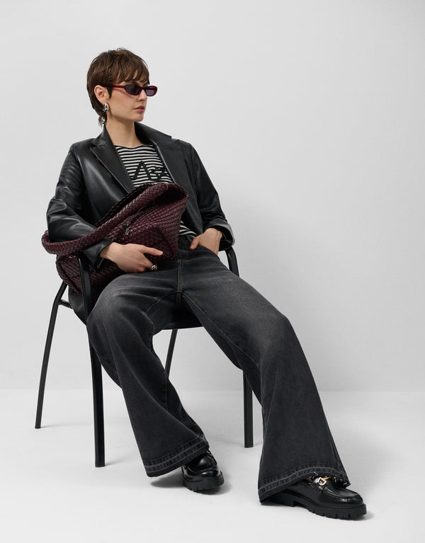 Person sitting on a chair wearing a black leather jacket, black pants, and sunglasses, holding a maroon clutch.