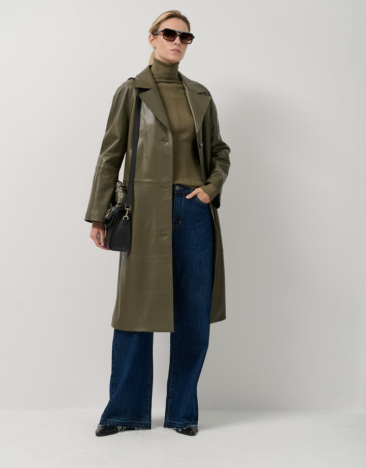 Woman wearing an olive green coat, turtleneck, and jeans on a white background