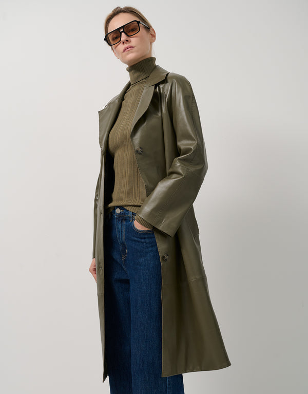 Person wearing a long green coat, brown turtleneck, and blue jeans on a white background