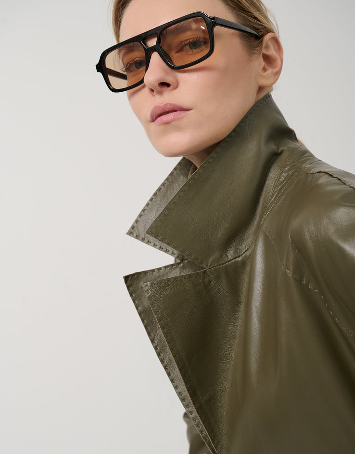 Person wearing a green leather jacket and sunglasses on a light gray background