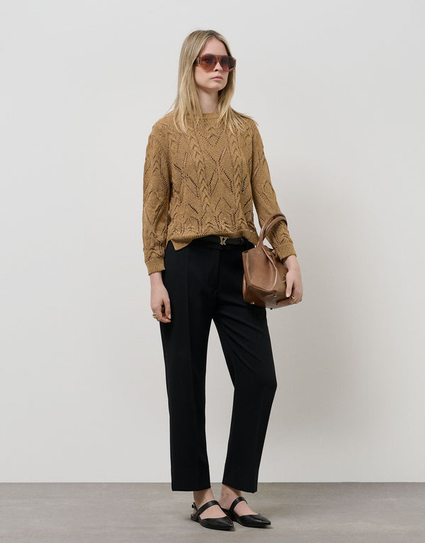 Woman wearing a beige sweater and black pants, holding a brown bag against a plain background