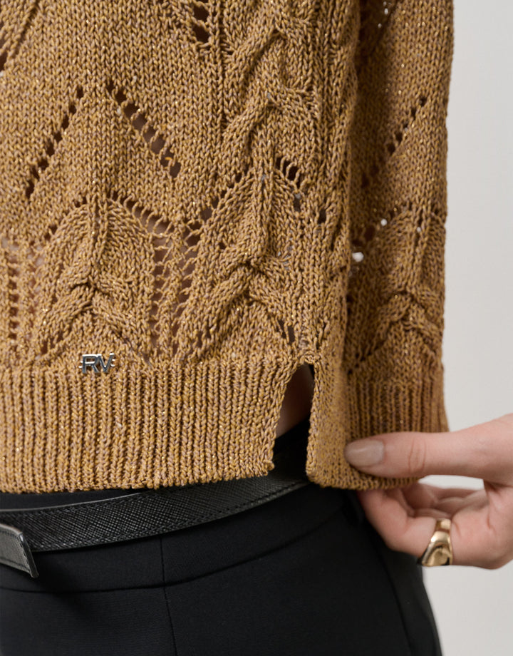 Close-up of a brown cable knit sweater with a brand logo, worn over black pants.
