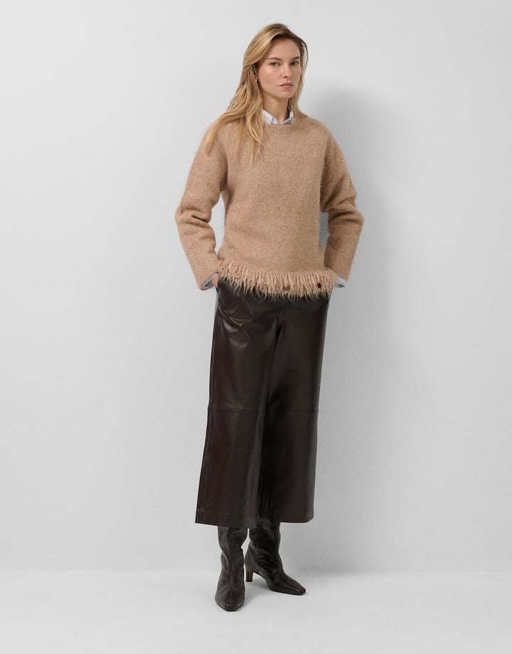 Woman wearing a beige sweater with fringe and black leather pants on a white background