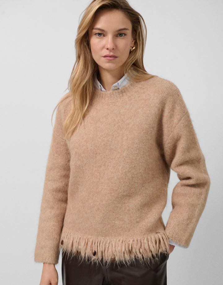 Woman wearing a beige sweater with fringe details on a plain background