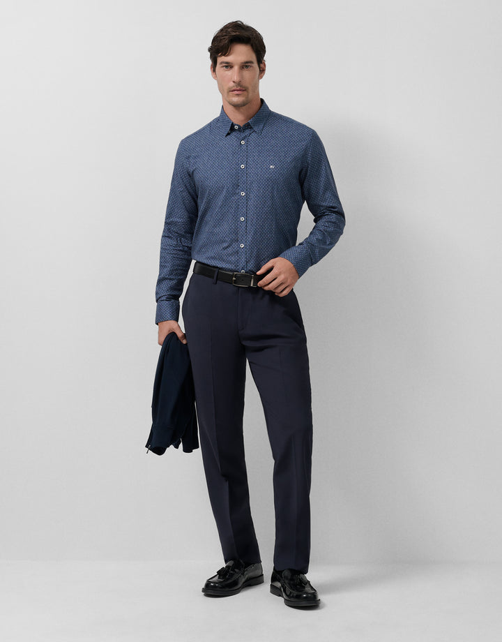 Man wearing a blue patterned shirt and dark pants on a light gray background