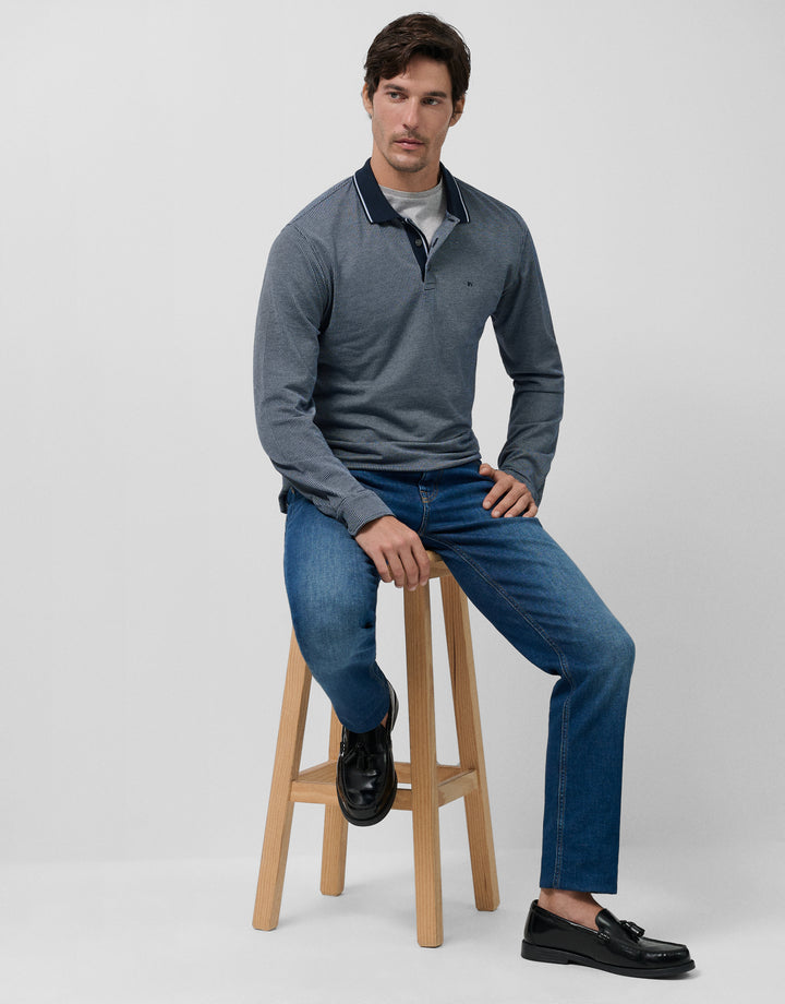 Man sitting on a wooden stool wearing a gray sweater, blue jeans, and black shoes against a white background