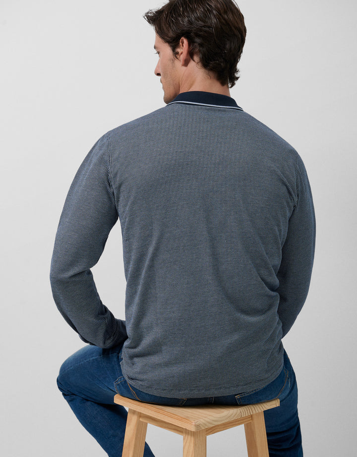 Man sitting on a stool wearing a gray long-sleeve shirt and blue jeans against a plain background