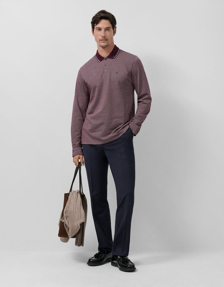 Man wearing a patterned long-sleeve shirt and dark pants, holding a jacket against a light gray background
