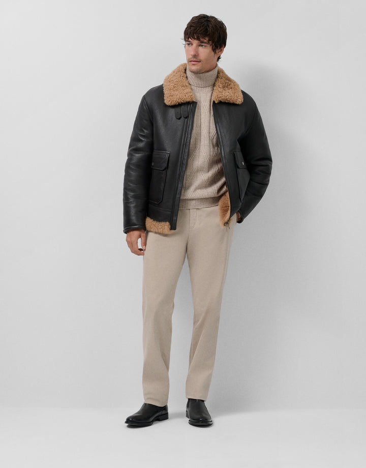 Man wearing a black leather jacket with fur collar, beige sweater, and pants on a white background