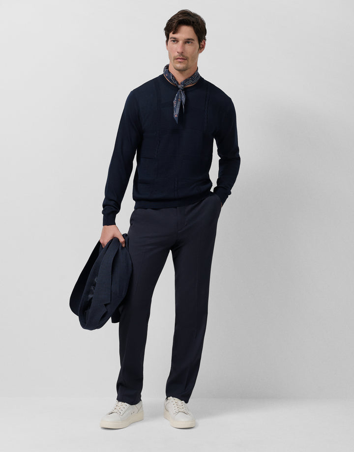 Man wearing a navy sweater and pants holding a jacket on a light gray background