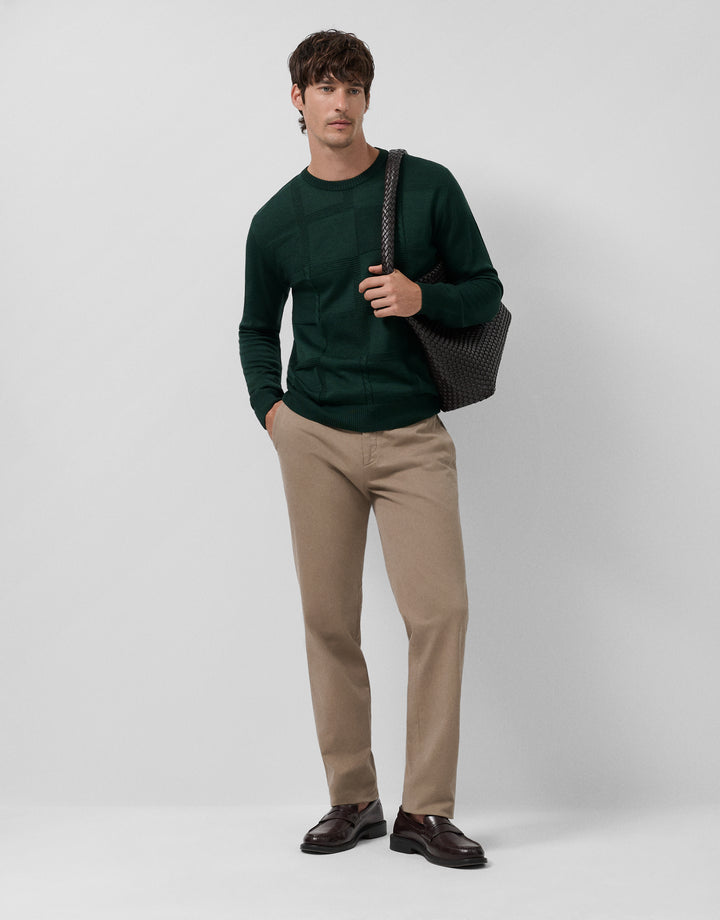 Man wearing a green sweater, beige pants, and brown shoes holding a black bag on a white background